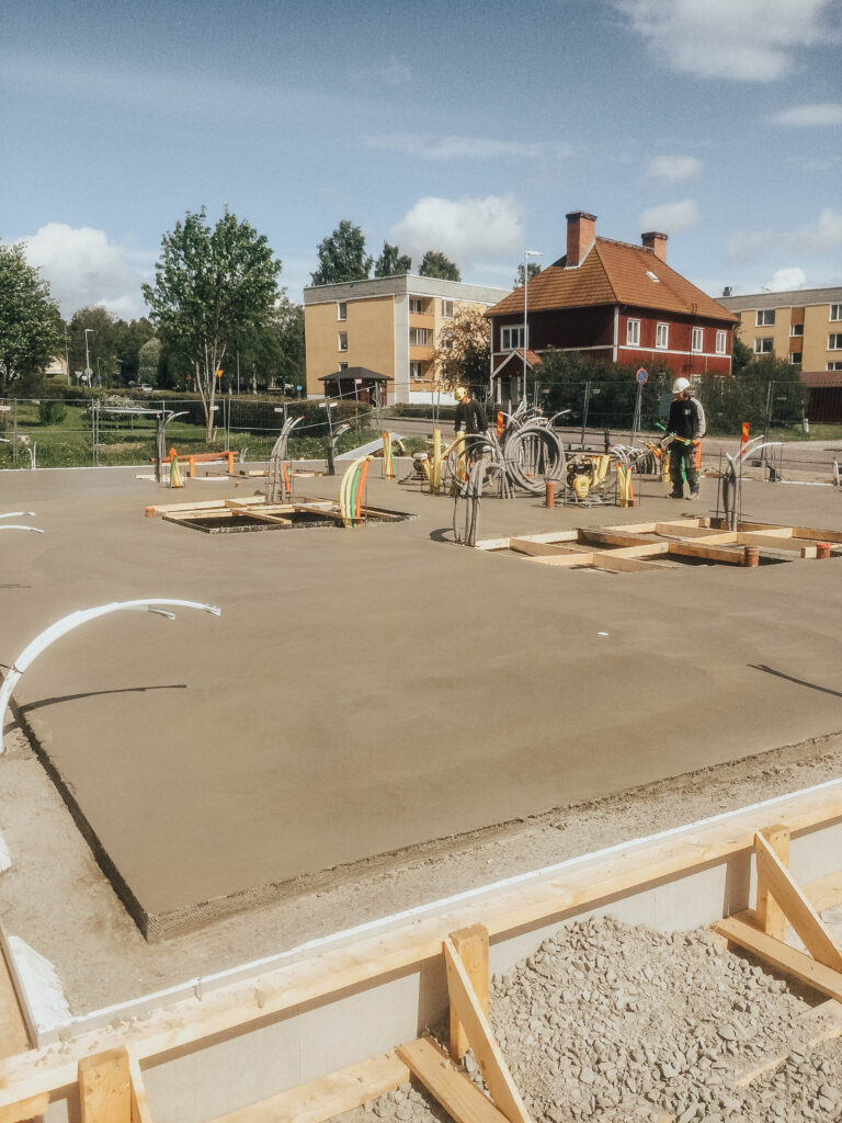 Två män i arbetskläder med glättare på betongplatta förberedd för husbygge. Rör sticker upp ur betongplattan på flera ställen.